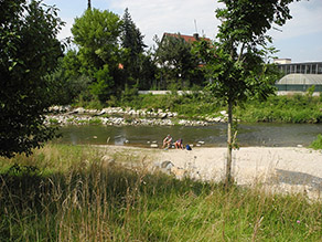 Zwei Personen sitzen auf Steinen am abgeflachten Kiesufer eines Flusses.