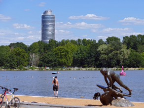 An einem Sandstrand steht eine Neptun-Statue und ein Fahrrad lehnt an einer niedrigen Sitzmauer. Dahinter steht ein Mann oberkörperfrei am Ufer eines aufgestauten Flusses auf dem sich Wasservögel und zwei Tretboote befinden. Am gegenüberliegenden Ufer ein Business Tower hinter Bäumen.