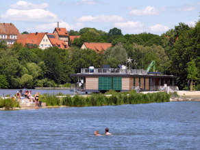 Zwei Personen schwimmen im Wasser. Im Hintergrund mehrere Personen auf Sitzsteinen am Ufer. Dahinter eine weitere Wasserfläche und die Energie- und Umweltstation Nürnberg.