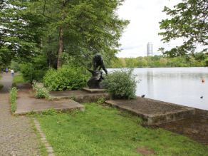 Eine Statue steht auf einem Betonsockel am Ufer eines Gewässers.