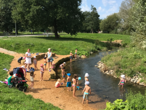 In der Außenkurve eines kleinen Bachs befindet sich ein Sandspielbereich mit flachen Ufern und zwei Sitzsteinmauern. Viele Kinder stehen am Ufer oder im flachen Wasser.