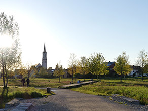 Auf einem Geh- und Radweg, der durch eine Grünanlage führt, spazieren zwei Personen in der Abendsonne. Auf einem Holzsteg auf einer Wiesenfläche sitzen mehrere Personen. Im Hintergrund Wohnbebauung und eine Kirche.