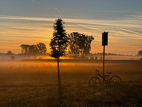 Nebelschwaden ziehen im orangefarbenen Licht der untergehenden Sonne über eine Wiesenlandschaft. Ein Fahrrad lehnt an einem Schild im Vordergrund.