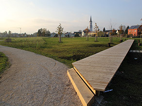 Ein Geh- und Radweg verläuft durch eine Grünanlage mit Wiesenflächen und zahlreichen Bäumen. Ein Holzsteg führt in die Wiese. Im Hintergrund Wohnbebauung und eine Kirche.