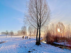 Drei Personen spazieren auf einem Weg parallel zu einem Bach durch eine winterliche Schneelandschaft bei Sonnenuntergang.