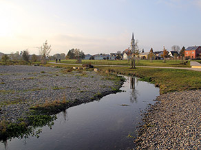 Parallel zu einem Bach mit abgeflachten, kiesigen Ufern verläuft ein Geh- und Radweg. Im Hintergrund eine Grünanlage mit Wiesenflächen und zahlreichen jungen Bäumen, sowie eine Siedlung mit Kirche.