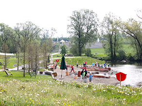Am Flachufer eines Flusses spielen zahlreiche Kinder an einem Wasserspielplatz. Bänke stehen auf einer Wiesenfläche daneben.