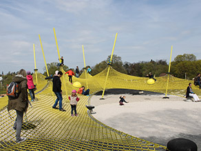 Auf einem Spielplatz mit Sandfläche und einem Gerüst aus Netzen spielen zahlreiche Kinder und Erwachsene.