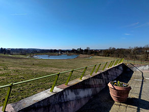 Blick über die Wilhelminenaue von einer Aussichtsplattform mit Geländer. Im Hintergrund die Grünanlage mit mehreren Wegen und ein Landschaftssee mit Seebühne.