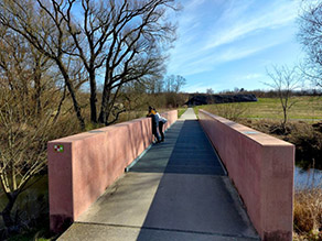 Auf einer Brücke über einen Fluss mit Betonmauern als Brüstung stehen zwei Personen und betrachten das Gewässer.