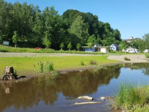 Ein Fluss mit sehr flachen Ufern und daran angrenzenden Kies- und Wiesenflächen. Im Hintergrund ein Wohnmobilstellplatz.