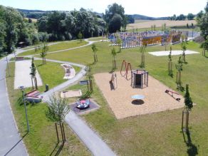Luftaufnahme einer Grünanlage mit Kinderspielplatz, einem Geh- und Radweg und Wiesenflächen mit vereinzelten jungen Bäumen. Im Hintergrund ein Schilderpark (Fernweh-Park).