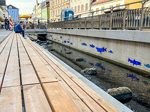 Sitzmöglichkeiten aus Holz am geöffneten Moosach-Kanal. Im Wasser ist ein kiesiges Bachbett mit Trittsteinen zu erkennen. Auf der gegenüberliegenden Kanalwand sind Fische aufgezeichnet.