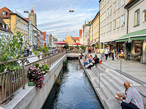 Durch den Stadtplatz an der Oberen Hauptstraße in Freising fließt der freigelegte Moosach-Kanal. Stufen führen vom Stadtplatzniveau hinab zum Wasser und bieten auch Sitzgelegenheiten, auf denen Menschen mit Eiscreme sitzen. Die gegenüberliegende Kanalseite ist mit Kübelpflanzen begrünt.