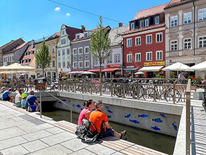 Blick auf die freigelegte Moosach am Freisinger Stadtplatz mit seinen Sitzstufen. Die gegenüberliegende Wand des Kanals ist bemalt mit Fischen. Einige Fahrradfahrer nutzen das Angebot für eine Rast am Wasser.