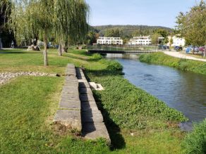 Am abgeflachten Ufer eines Flusses zwei Sitzreihen aus Steinquadern. Weiden wachsen entlang des Ufers. Im Hintergrund eine Fußgängerbrücke über den Fluss.
