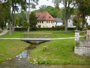 Der Webbach (Seitengewässer der Sulz) fließt durch eine Grünanlage mit Fußgängerbrücke und mündet in einen Fluss. Im Hintergrund Wohnbebauung.