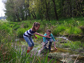 Zwei Kinder stehen mit Gummistiefeln in einem flachen Bach und spritzen mit Wasser um sich. Im Hintergrund windet sich der Bach durch hohes Gras.