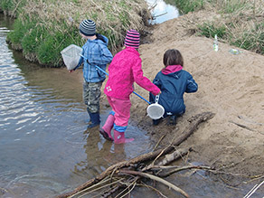 Drei Kinder mit Gummistiefeln stehen und hocken an einem Bach mit sandigem Ufer und untersuchen das Gewässer mit Keschern.