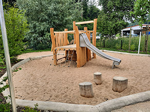 Neuer Spielplatz: Ein Klettergerüst aus Holz mit Rutsche und drei Holzstempen befinden auf einer Sandfläche eingefasst von mehreren Baumstämmen.