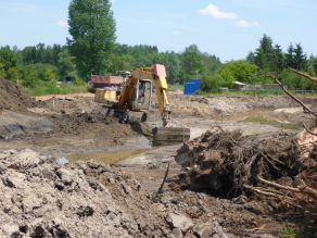 Umgeben von Erdhaufen und Wurzelstöcke gräbt ein Bagger gräbt mit einer Schaufel das neue Gewässerprofil aus, welches bereits teilweise mit Wasser gefüllt ist.