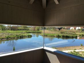 Blick aus dem Innern eines Pavillons durch ein Sichtfenster auf eine Gewässerlandschaft. Im Hintergrund eine grasbewachsene Insel, die von Wasser umgeben ist.
