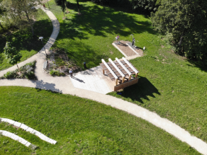 Luftaufnahme der neuen Parkanlage, die den neu angelegten Wegeverlauf, eine Sitztribüne, einen Pavillon und zwei Fitnessgeräte zeigt. Umgeben ist die Anlage von grünen Bäumen.