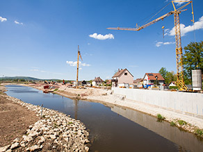 Neben dem neuen Gewässerlauf wird eine Ufermauer hergestellt. Zwei Turmdrehkräne stehen am Ufer.