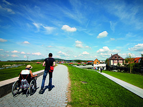 Eine gehende und eine rollstuhlfahrende Person auf einem Geh- und Radweg. Seitlich ist der Gewässerlauf der Wörnitz zu erkennen.