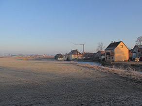 An einen Fluss im Bereich des Unterwassers einer Mühle grenzt eine Grünlandfläche. Im Hintergrund eine Siedlung mit Wohnbebauung.