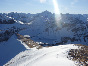 Mitte Januar hat starker Wind den Schnee im Lee abgelagert. Die windzugewandten Hangbereiche sind bis zu Boden abgeblasen. Im Hintergrund sind die Gipfel der Allgäuer Hochalpen vor wolkenlosem Himmel zu sehen.