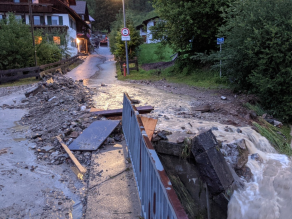 Wasser läuft zwischen Häusern eine schmale Straße hinunter und fließt in einen Bach, der einen erhöhten Abfluss hat. Auf der Straße wurden Geröll, Bodenmaterial, Holzplatten und Unrat angeschwemmt.