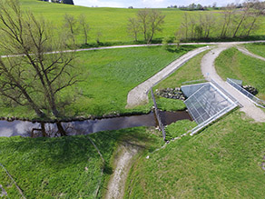 Foto eines Hochwasserrückhaltebeckens in Thalhofen. Zu sehen ist ein Weg der über den Staudamm führt, das Gewässer bei normalem Wasserstand sowie die Betriebseinrichtungen.